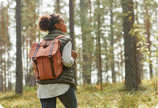 Back view portrait of young African-American woman with backpack enjoying hiking in forest lit by sunlight