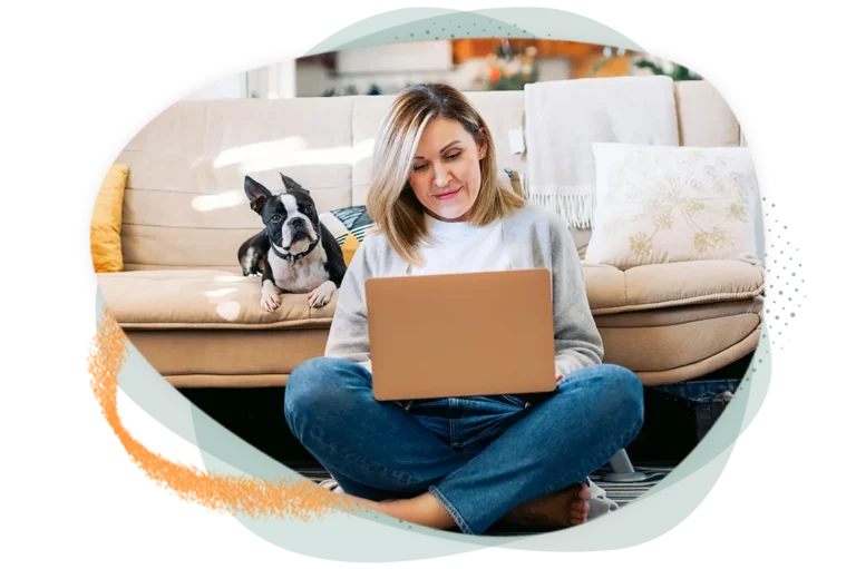 Woman sitting in front of couch on her laptop with her dog in the background