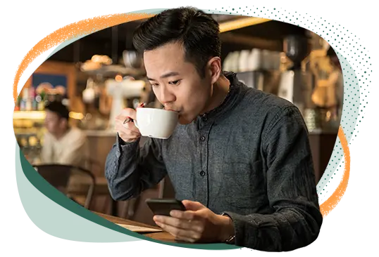 Asian male sitting at a table sipping coffee and looking at his phone.